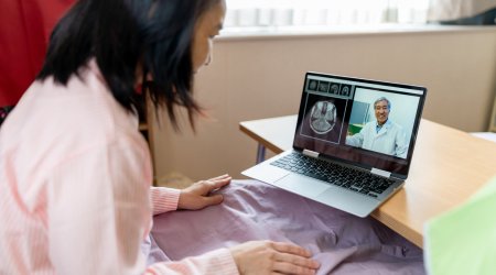Sick Japanese woman on bed video chatting with doctor on laptop at home. The image on the left side of the monitor screen is the head image obtained by the MRI scan.